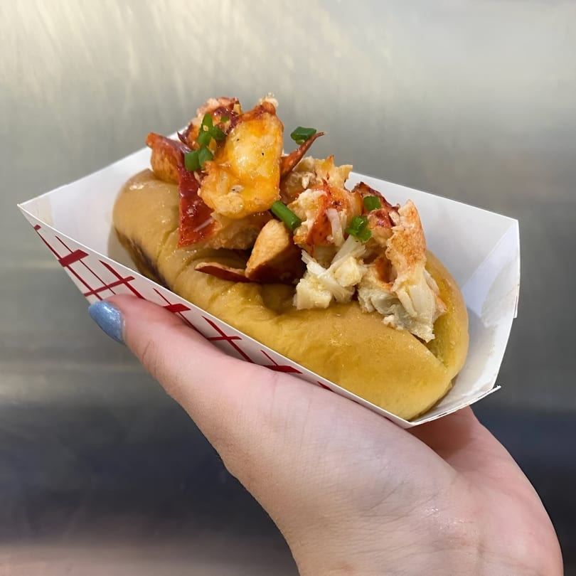 A hand holds a Boca Lobster Roll in a paper tray, filled with lobster meat and green onions.