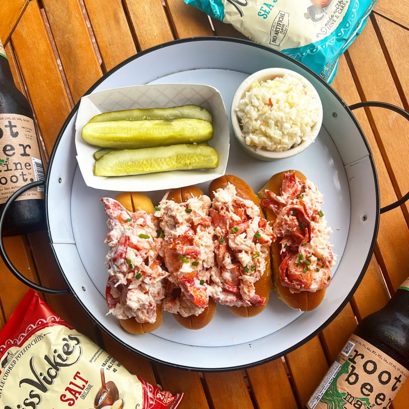 Four Boca Lobster Rolls with pickles and coleslaw on a white tray, with root beer and chips.