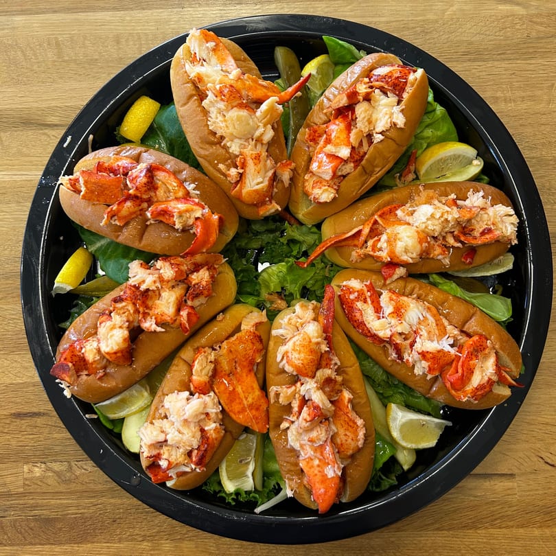 Eight Boca Lobster Roll Company lobster rolls arranged on a black platter with lettuce and lemon wedges.