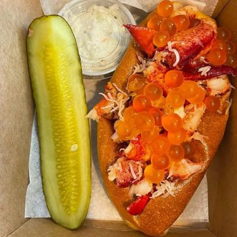 A Boca Lobster Roll with orange roe, a pickle spear, and coleslaw in a takeout container.