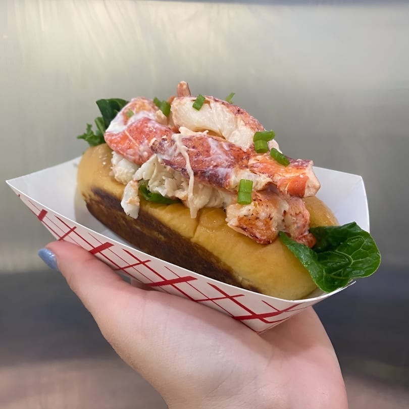A hand holds a Boca Lobster Roll in a paper tray, filled with lobster, lettuce, and chives.