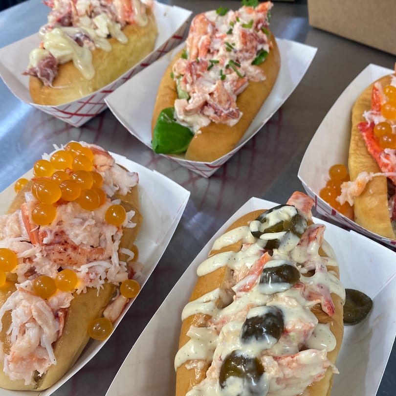 Four Boca Lobster Roll Company lobster rolls with various toppings in white paper trays.