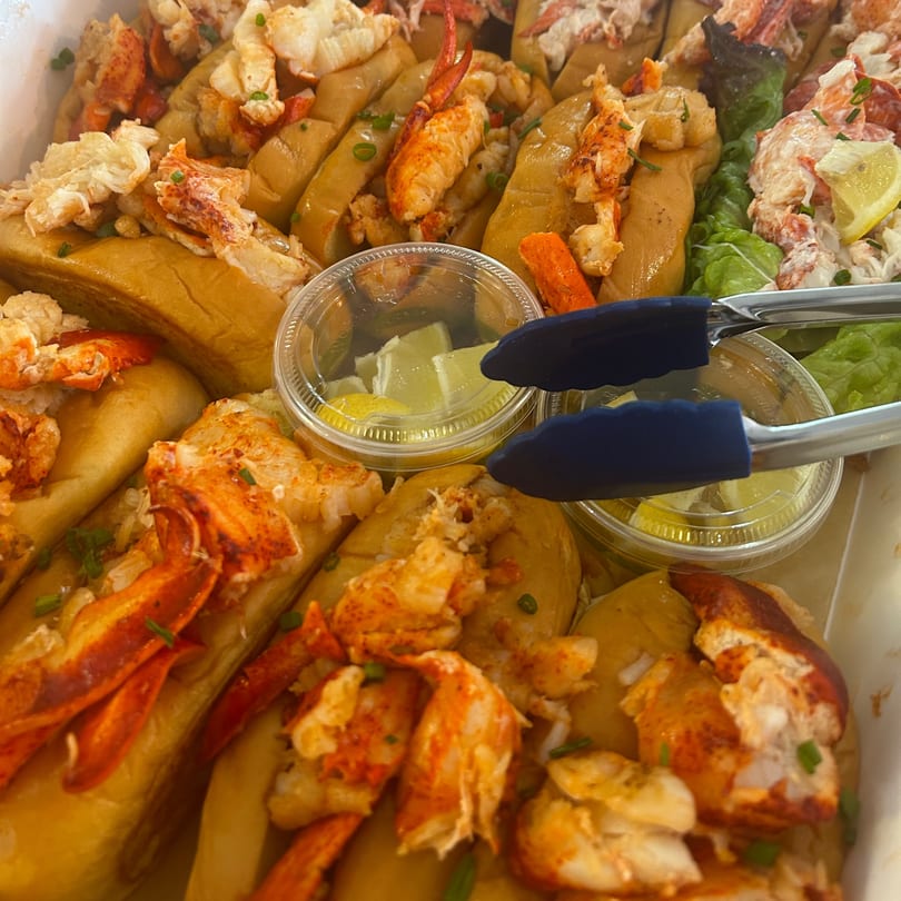 A tray of Boca Lobster Roll Company's lobster rolls, with lemon wedges and tongs.
