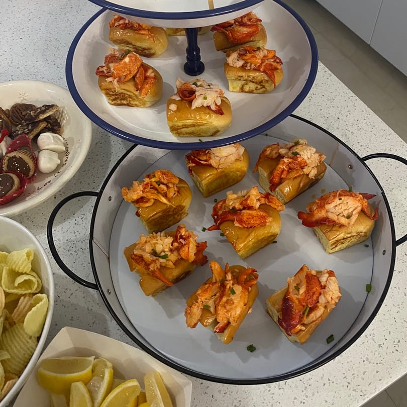 A two-tiered tray of mini lobster rolls, with chips, lemon wedges, and desserts on a counter.